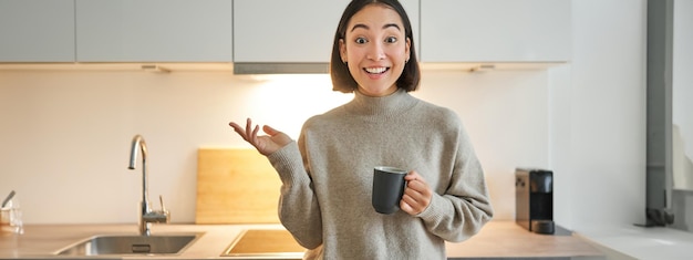 Free photo portrait of stylish asian woman standing in kitchen with mug drinking coffee explaining smth showing