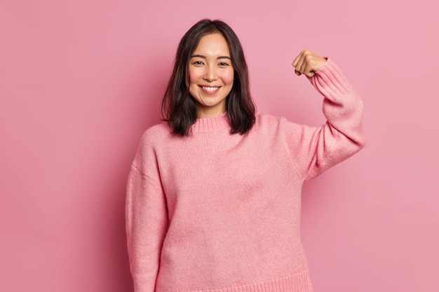 Free Photo powerful cheerful brunette woman raises arm and shows muscle demonstrates her strength looks confident at camera smiles gently wears casual long sleeved jumper 