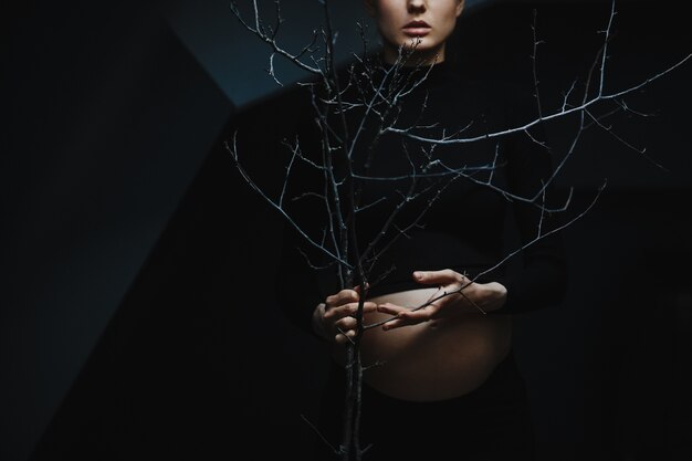 Pregnant woman in black clothes stands under grey wall and holds a tree branch 