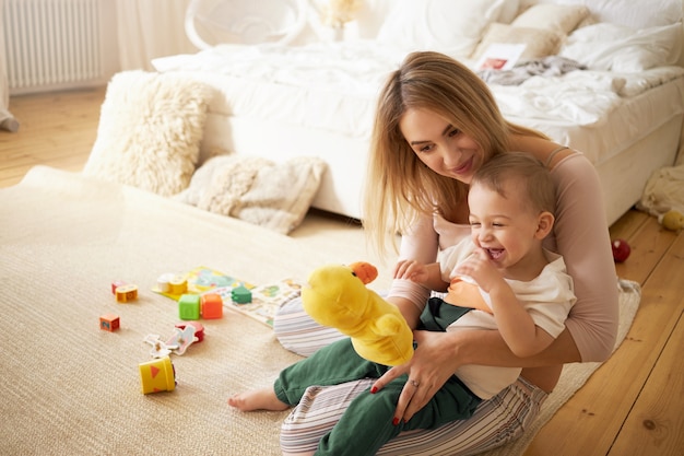 Free Photo pretty sister spending time with her baby brother, sitting on floor in bedroom. beautiful young babysitter playing with little boy indoors, holding stuffed toy duck. infancy, childcare and motherhood