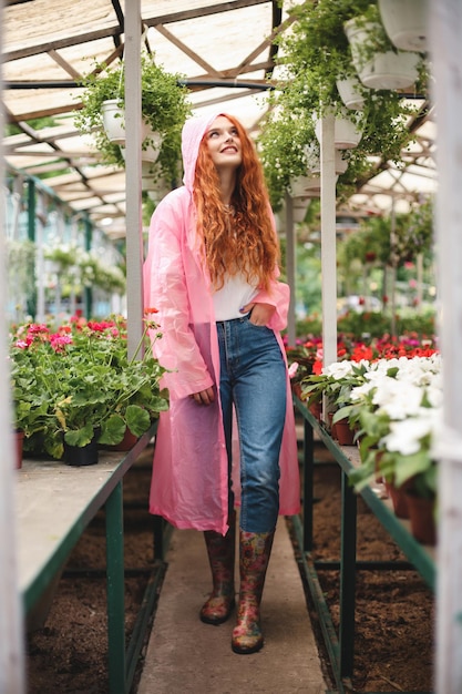 Free photo pretty smiling lady with redhead curly hair walking in pink raincoat and happily aside while spending time in greenhouse