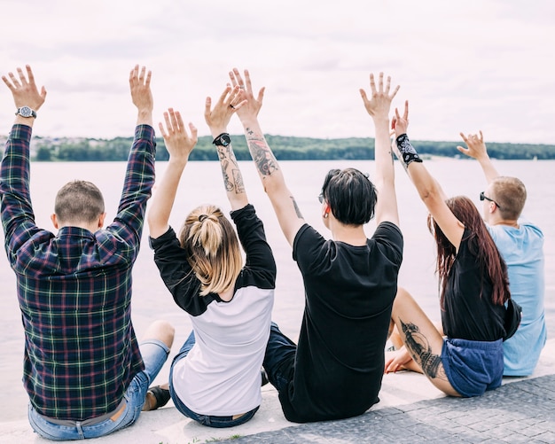 Free photo rear view of friends sitting near the lake raising their hands