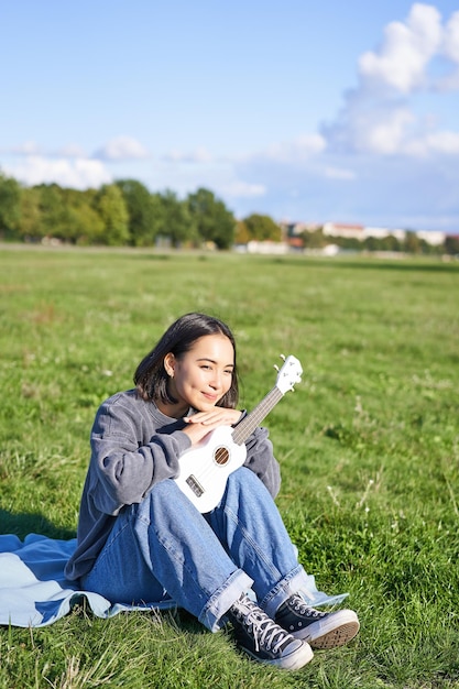 Romantic asian girl sitting with ukulele guitar in park and smiling relaxing after university enjoyi