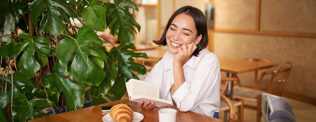 Бесплатное фото Романтическая азиатка сидит с книгой в кафе, ест круассан и пьет кофе, читает и