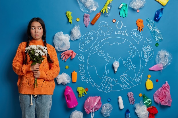 Free photo sad disappointed nature preserver stands with bouquet against blue background with drawn planet and light bubble in center, thinks about saving world from contamination.