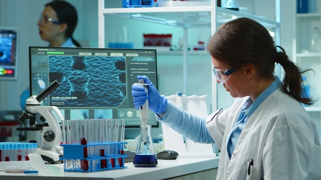 Scientist woman pipetting liquid to test tube late at night in modern equipped lab. Team examining virus evolution using high tech for scientific analysis of treatment development against covid19