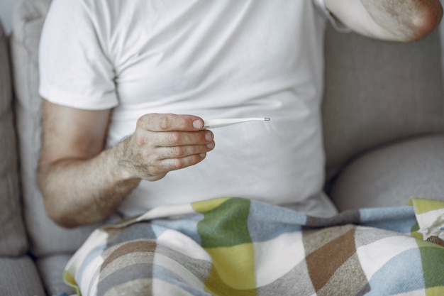 Free photo senior man alone sitting on sofa. sick man covered with plaid. grangfather with thermometer.