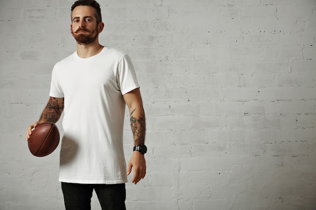 Foto gratuita giovane attraente serio in una maglietta di cotone bianca vuota e jeans neri che tengono un pallone da rugby vintage isolato su bianco