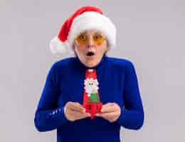 Free photo shocked elderly woman in sun glasses with santa hat and santa tie holding christmas tree ornament