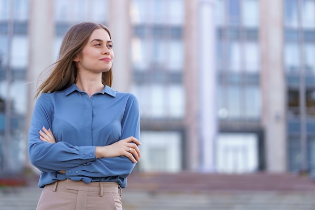 Shot of beautiful young businesswoman wearing blue chiffon shirt while standing on building in the street with folded arms.