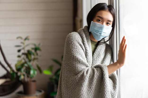 Free Photo sick asian girl in medical face mask standing by the window and yearning to go outside being on quar...