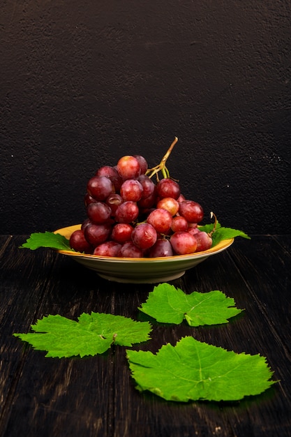 Free photo side view of a bunch of sweet grapes in a plate and green grape leaves on rustic table