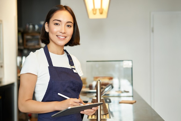 Foto gratuita barista sorridente della ragazza asiatica di affari del piccolo caffè in grembiule facendo uso della compressa come coff di elaborazione del terminale di posizione