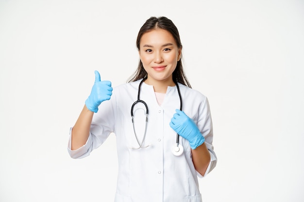 Foto gratuita la dottoressa asiatica sorridente mostra i pollici in su, indossa guanti di gomma e uniforme clinica, si erge su sfondo bianco