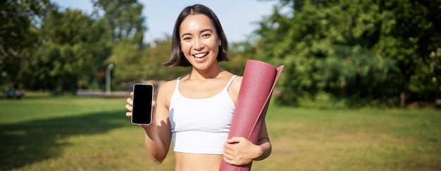 Free photo smiling asian fitness girl with rubber yoga mat shows her smartphone screen recommends workout