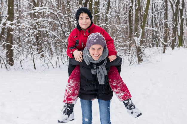 Free photo smiling boy enjoying piggy back with his mother in winter at forest