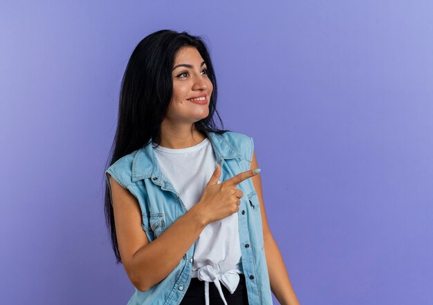 Smiling young caucasian girl looks and points at side isolated on purple background with copy space