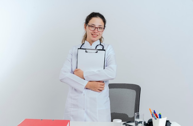 Foto gratuita sorridente giovane medico femminile che indossa abito medico e stetoscopio e occhiali in piedi dietro lo scrittorio con strumenti medici che sembrano appunti della holding isolati