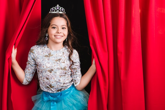 Free photo smiling young girl wearing crown looking out from red curtain