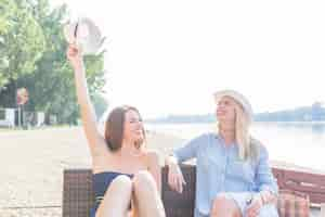 Free photo smiling young woman sitting with friends holding hat at beach