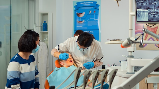 Foto gratuita stomatologo che parla con la madre dei bambini, accende la lampada ed esamina la bambina in piedi vicino alla sedia stomatologica. dentista pediatrico che parla con una donna mentre l'infermiera prepara gli strumenti sterilizzati.