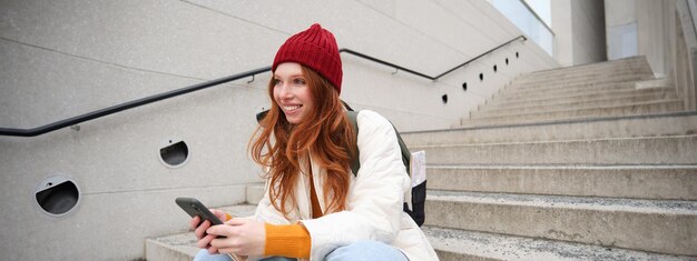 Free photo stylish european girl with red hair sits on public stairs with smartphone places online order sends