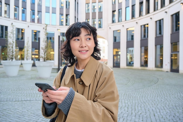 Free photo stylish korean girl in headphones listens music and uses mobile phone stands in city centre waits fo