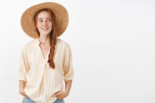 Free photo stylish redhead woman going to beach wearing straw hat not to tan turning right with happy carefree expression holding hands in pockets enjoying warm sunny summer day
