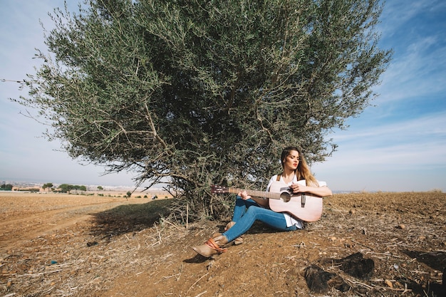 Foto gratuita donna alla moda che gioca chitarra vicino al cespuglio