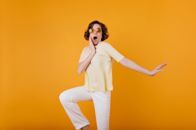 Free Photo surprised white woman in trendy yellow glasses standing on bright wall. shocked girl with wavy brown hair touching her face while posing .