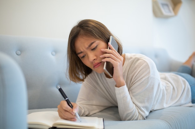 Free photo tensed lady making notes and calling on phone on sofa