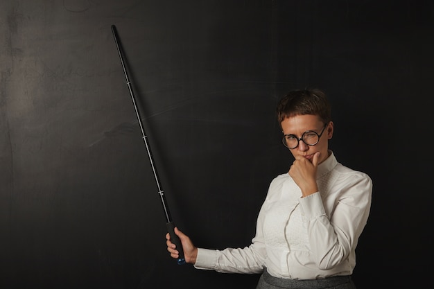 Thinkful business woman in front of school chalk board shows something with folding pointer and looks on side