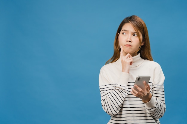 Free photo thinking dreaming young asia lady using phone with positive expression, dressed in casual clothing feeling happiness and stand isolated on blue wall