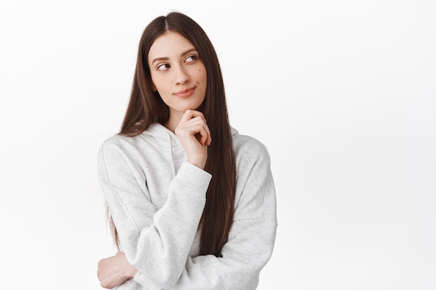 Free photo thoughtful cute girl with long hair looking aside and smiling while thinking, standing pensive, making choice, imagine something interesting, white wall