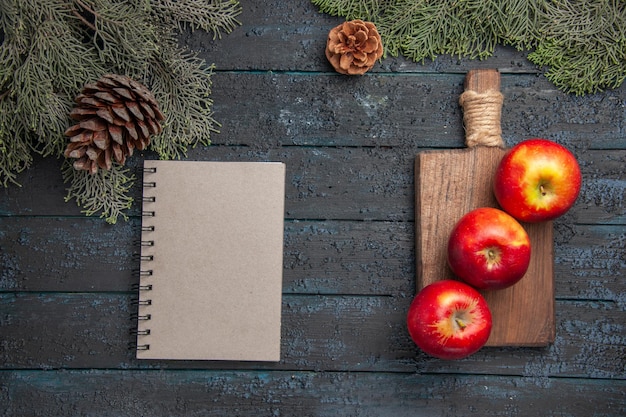 Free photo top view apples on board three apples on wooden cutting board and notebook under branches with cones