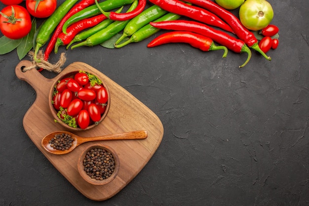 Top view hot red and green peppers and tomatoes bay leaves bowls with cherry tomatoes and black pepper and spoon on a chopping board on black ground with free space