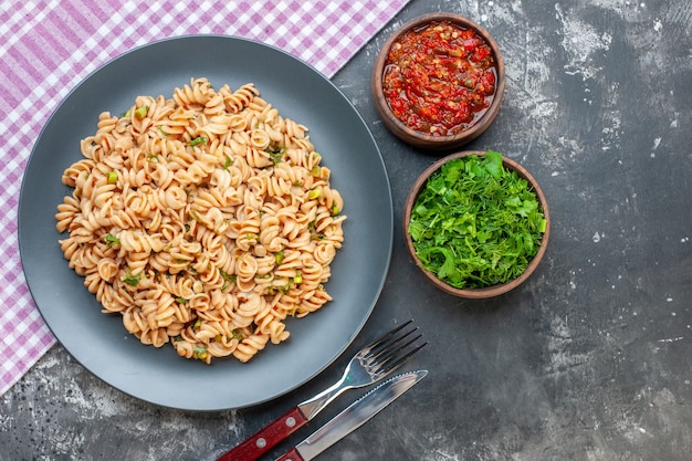 Foto gratuita pasta rotini vista dall'alto su piatto rotondo su salsa di pomodoro tovaglia a scacchi bianco rosa e verdure tritate in piccole ciotole coltello e forchetta su superficie scura