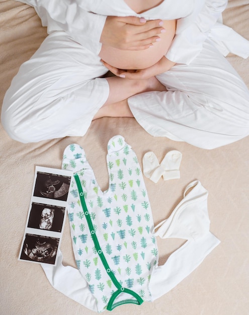 Free photo above view of anonymous woman in white cozy pajama sitting on bed with ultrasound image of womb cute bodysuit and socks while caressing pregnant belly and expecting for baby and part of family