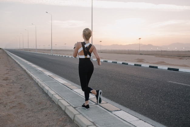 Free photo view from back workout on road of attractive young woman in sportswear running in sunny morning.healthy lifestyle, training, strong sportswoman.