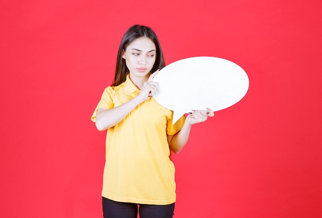 Foto gratuita donna in camicia gialla che tiene una scheda informativa ovale e sembra confusa e pensierosa.