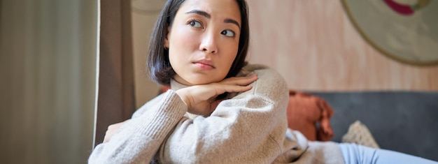 Free photo women and wellbeing concept portrait of thoughtful asian woman sitting on sofa with pillow looking