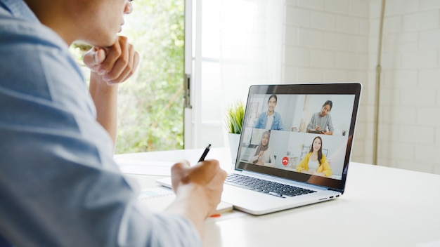 Free Photo young asia businessman using laptop talk to colleagues about plan in video call meeting while work from home at living room. self-isolation, social distancing, quarantine for corona virus prevention.