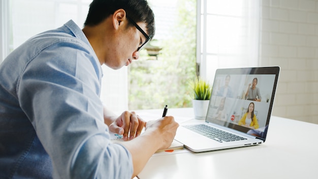 Free Photo young asia businessman using laptop talk to colleagues about plan in video call meeting while work from home at living room. self-isolation, social distancing, quarantine for corona virus prevention.