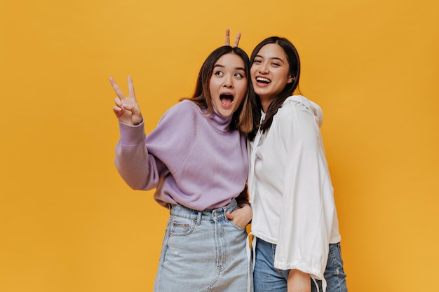 Free photo young asian girls laugh and pose on orange background pretty brunette woman in purple sweater shows peace sign goodhumored girl in white hoodie gives bunny ears to friend