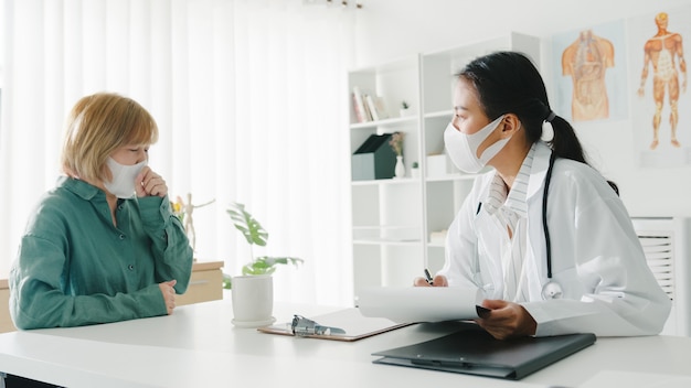 Free photo young asian lady doctor wearing protective mask using clipboard discussing results or symptoms with girl patient in hospital office.