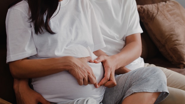 Free photo young asian pregnant couple making heart sign holding belly. mom and dad feeling happy smiling peaceful while take care baby, pregnancy lying on sofa in living room at home .