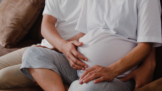 Free photo young asian pregnant couple man touch his wife belly talking with his child. mom and dad feeling happy smiling peaceful while take care baby, pregnancy lying on sofa in living room at home .