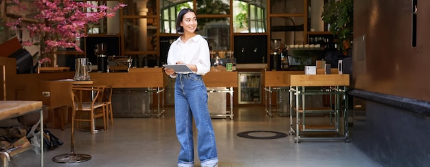 Free photo young asian woman with tablet standing in front of cafe entrance inviting guests and customers