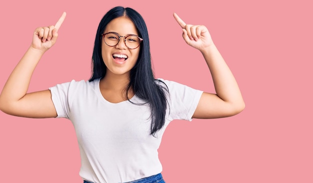 Free Photo young beautiful asian girl wearing casual clothes and glasses smiling amazed and surprised and pointing up with fingers and raised arms.