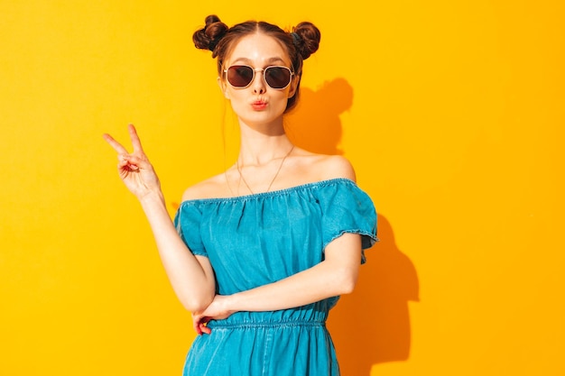 Free Photo young beautiful smiling female in trendy summer jeans overalls sexy woman with two horns hairstyle posing near yellow wall in studio positive model having fun shows peace sign in sunglasses